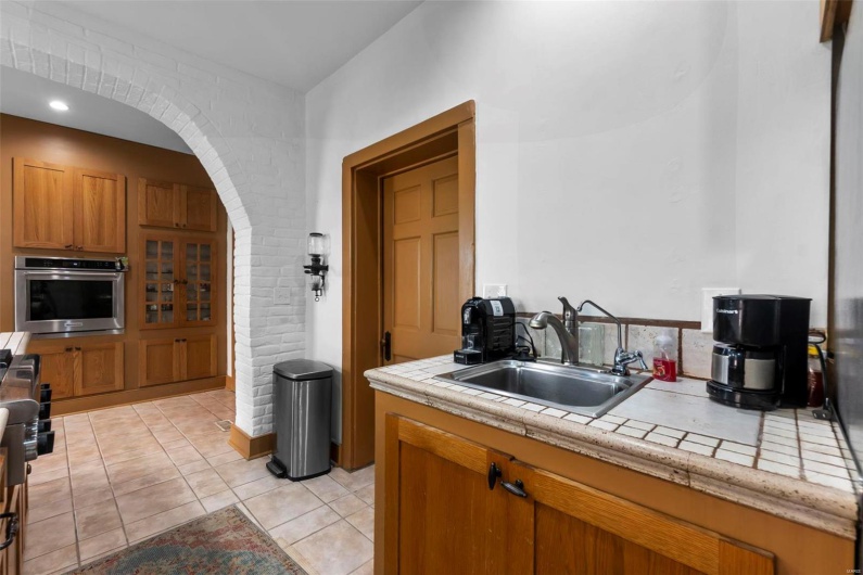 Kitchen featuring light tile patterned floors, stainless steel oven, tile counters, sink, and brick wall