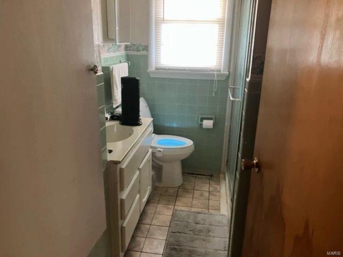 Bathroom featuring tile patterned flooring, toilet, a shower with door, vanity, and tile walls