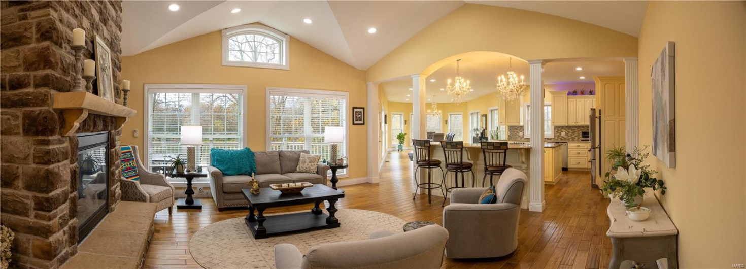Living room with ornate columns, high vaulted ceiling, a notable chandelier, a fireplace, and light hardwood flooring.