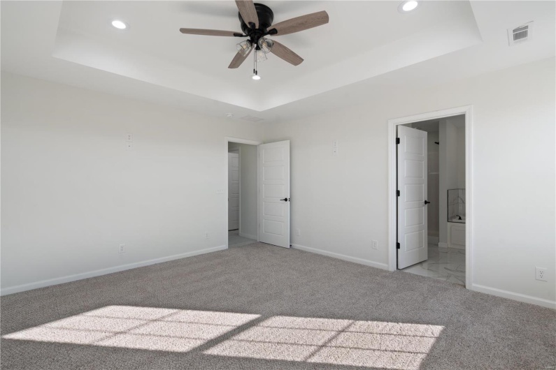 Unfurnished bedroom with ensuite bathroom, light colored carpet, ceiling fan, and a tray ceiling