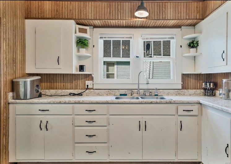 Kitchen with hanging light fixtures, sink, wooden walls, light stone counters, and white cabinetry