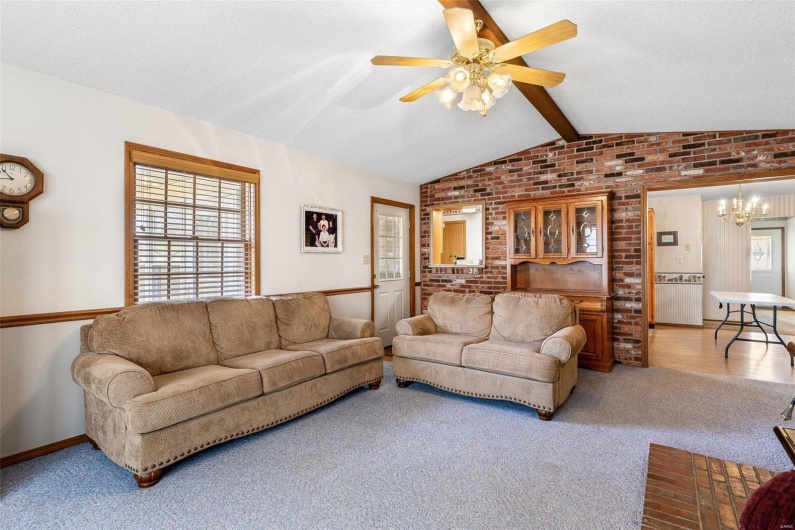 Living room with brick wall, vaulted ceiling with beams, a textured ceiling, carpet flooring, and ceiling fan with notable chandelier