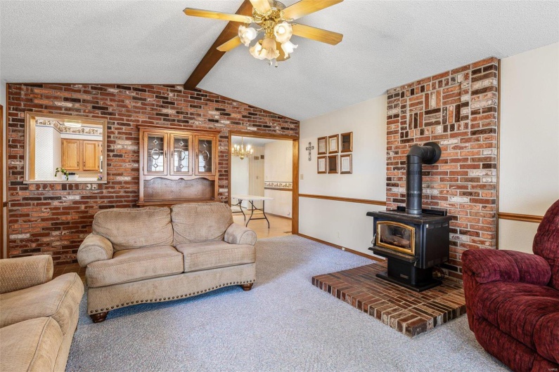Carpeted living area featuring lofted ceiling with beams, a textured ceiling, brick wall, ceiling fan, and a wood stove