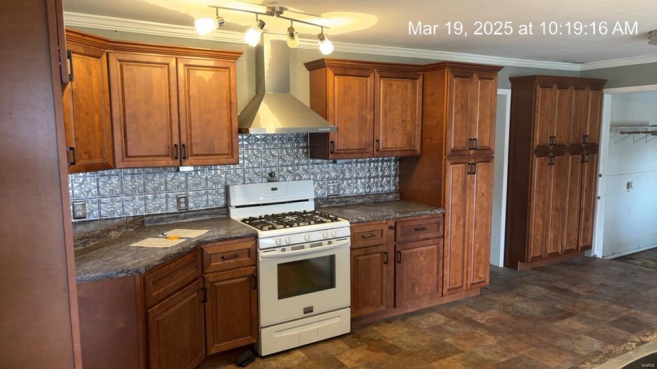 Kitchen featuring backsplash, white gas range, brown cabinetry, and wall chimney range hood