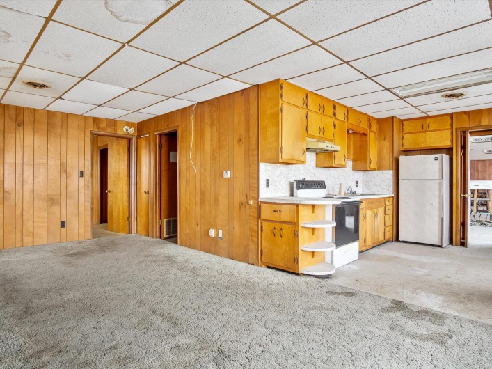 Kitchen featuring a paneled ceiling, white appliances, light colored carpet, sink, and wood walls