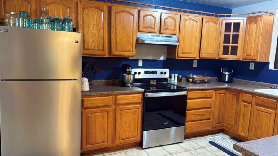 Kitchen featuring under cabinet range hood, brown cabinetry, stainless steel appliances, and glass insert cabinets