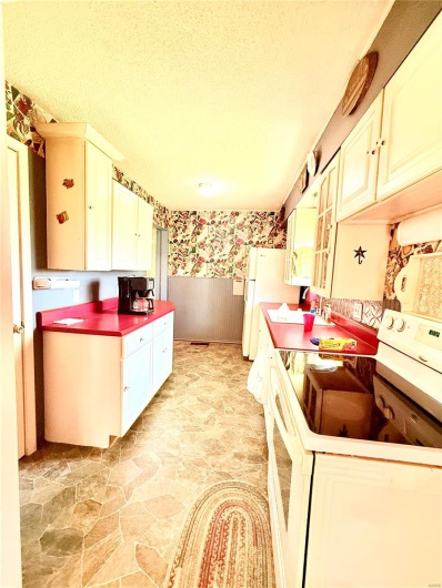 Kitchen featuring stone flooring, wallpapered walls, white appliances, wainscoting, and a textured ceiling