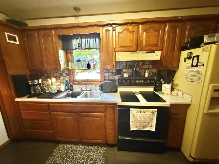 Kitchen with a sink, white refrigerator with ice dispenser, light countertops, under cabinet range hood, and black range with electric stovetop