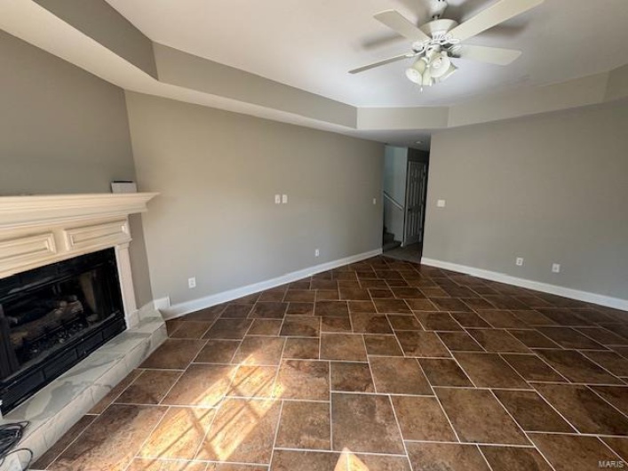 Unfurnished living room featuring baseboards, a fireplace with raised hearth, ceiling fan, and a tray ceiling