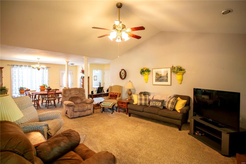 Carpeted living area featuring vaulted ceiling, decorative columns, and ceiling fan with notable chandelier