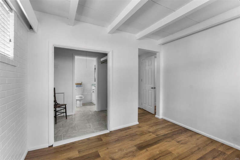 Unfurnished bedroom featuring beamed ceiling, wood finished floors, ensuite bathroom, baseboards, and brick wall