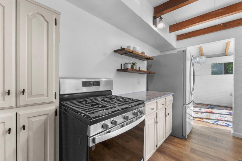 Kitchen with stainless steel appliances, light wood-type flooring, light stone countertops, white cabinetry, and beamed ceiling