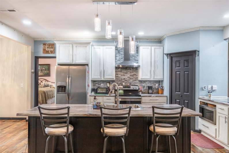 Kitchen with appliances with stainless steel finishes, wall chimney exhaust hood, decorative backsplash, an island with sink, and decorative light fixtures